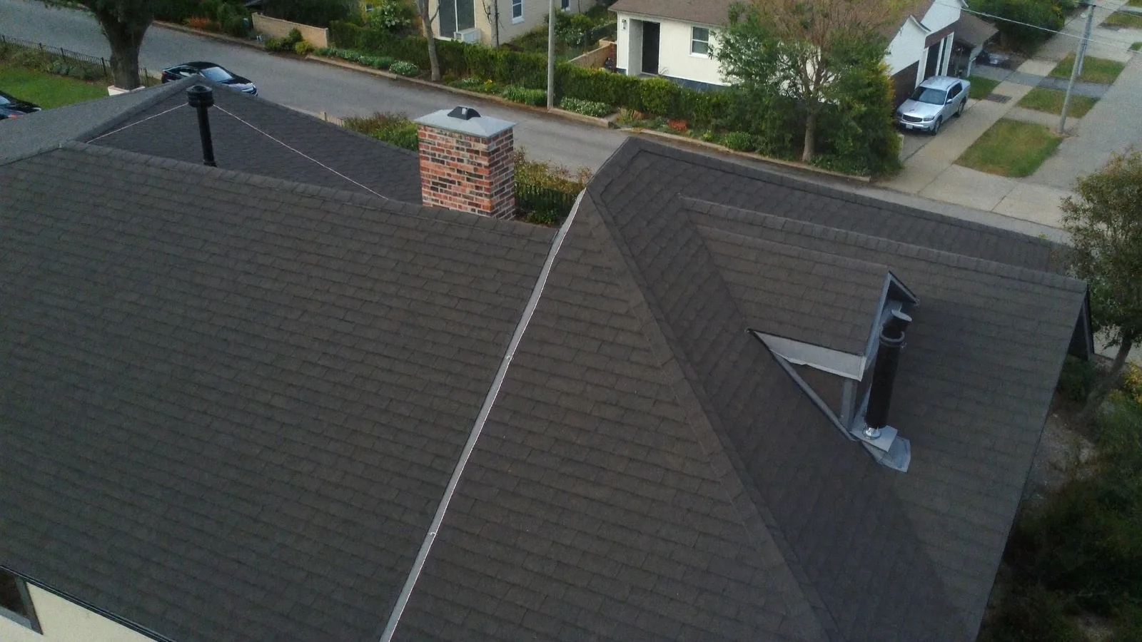 A drone view down onto a grey architectural shingle roof, showing two intersecting gable planes, a capped brick chimney and a flue on the rear slope