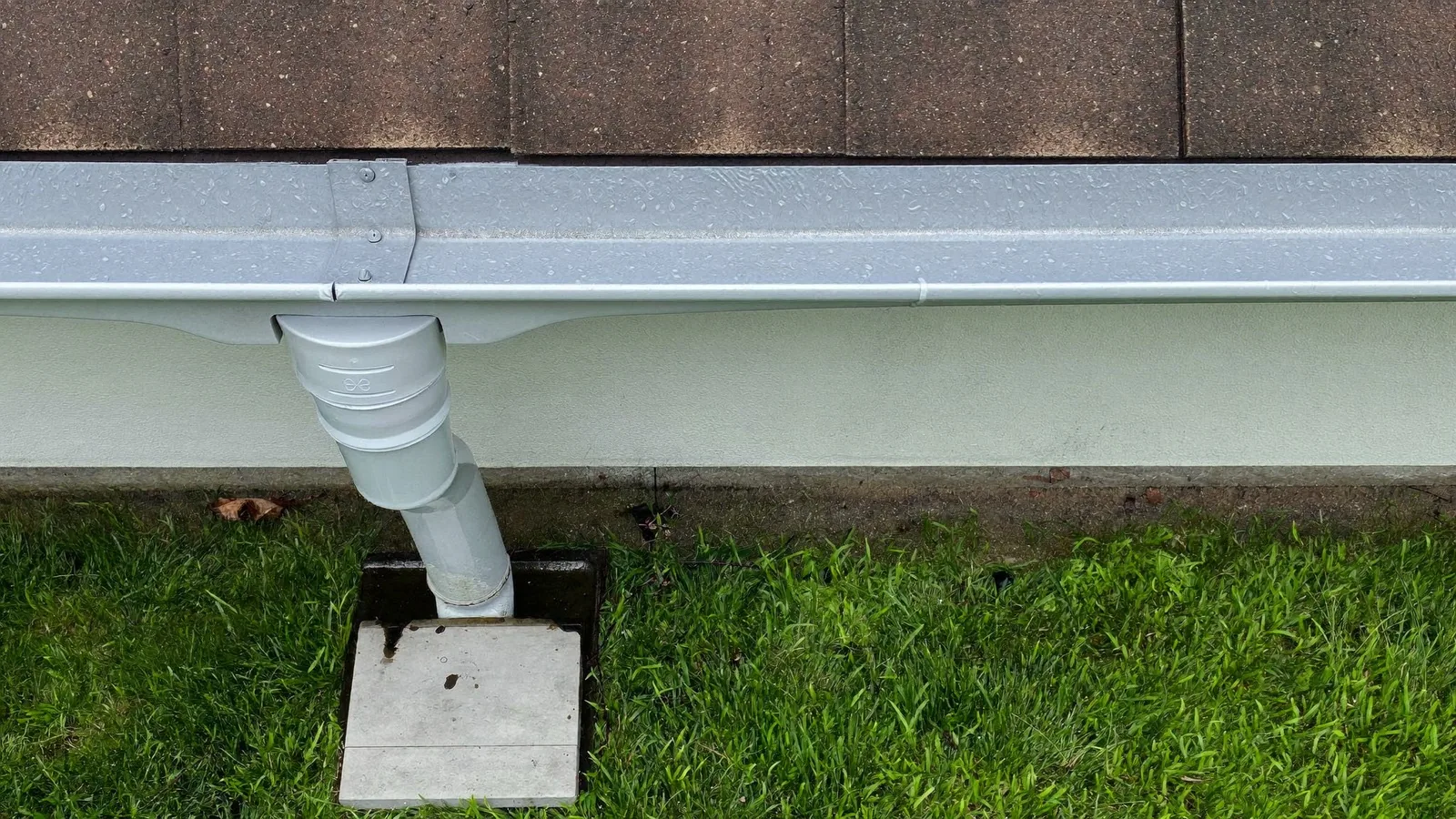 An aluminum gutter along a shingled eave, its downspout elbowing into a below-grade drain box set in the lawn