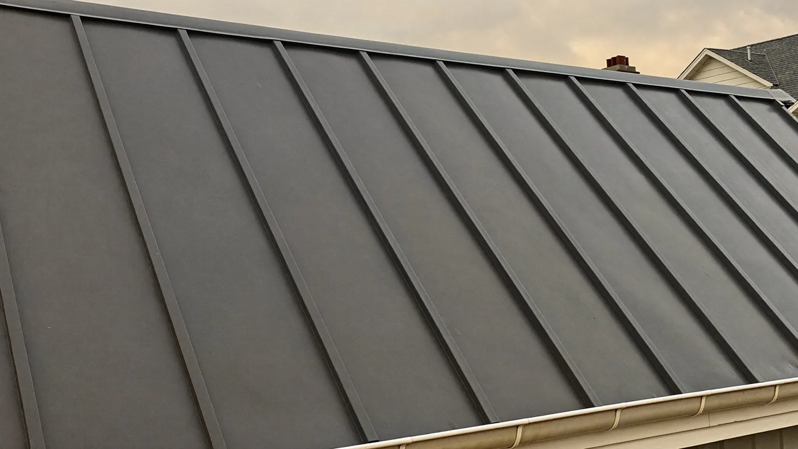 A dark grey standing-seam metal roof slope, the raised seams running up to the ridge cap, with a gutter along the eave