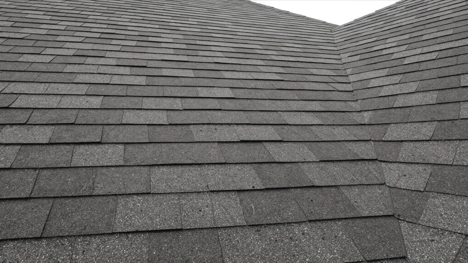 Looking up a grey shingle roof slope towards the ridge, the courses running away in even rows against a flat white sky